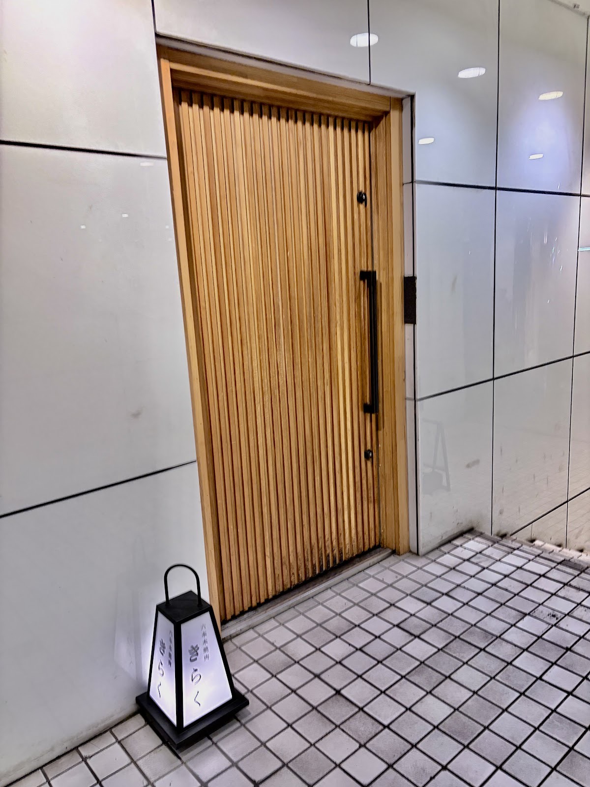 白い壁の通路にある木の縦格子の扉。足元に「六本木焼肉 きらく」と書かれた行灯。