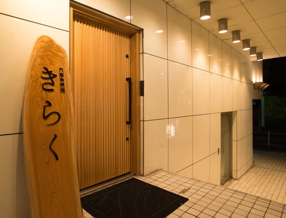 木の扉の横に立つ「六本木焼肉 きらく」の木の看板。夜の入口。