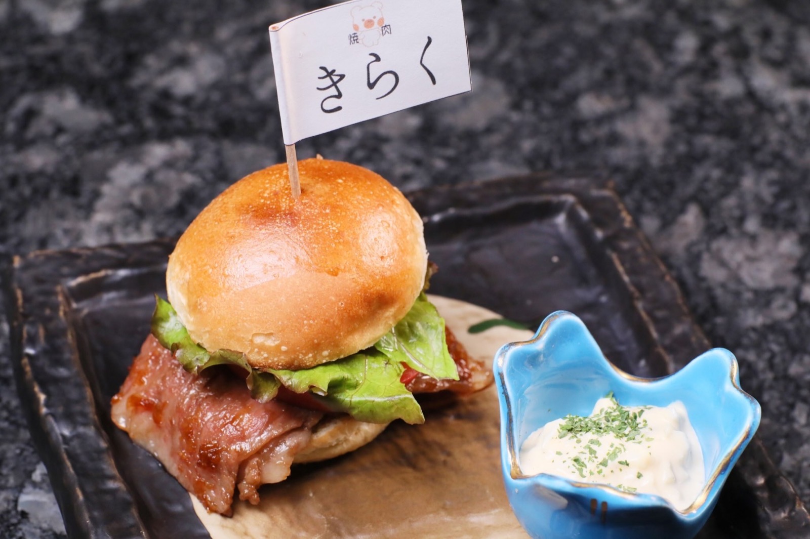 焼いた牛肉、レタス、トマトをはさんだ小さなバーガー。くまの顔と「焼肉 きらく」の小旗を立て、青い器に白いソース。