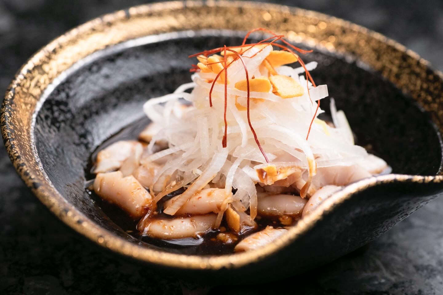 金色の縁の黒い器に、たれと白い切り身。細切りの白い野菜、ガーリックチップ、糸唐辛子をのせて。