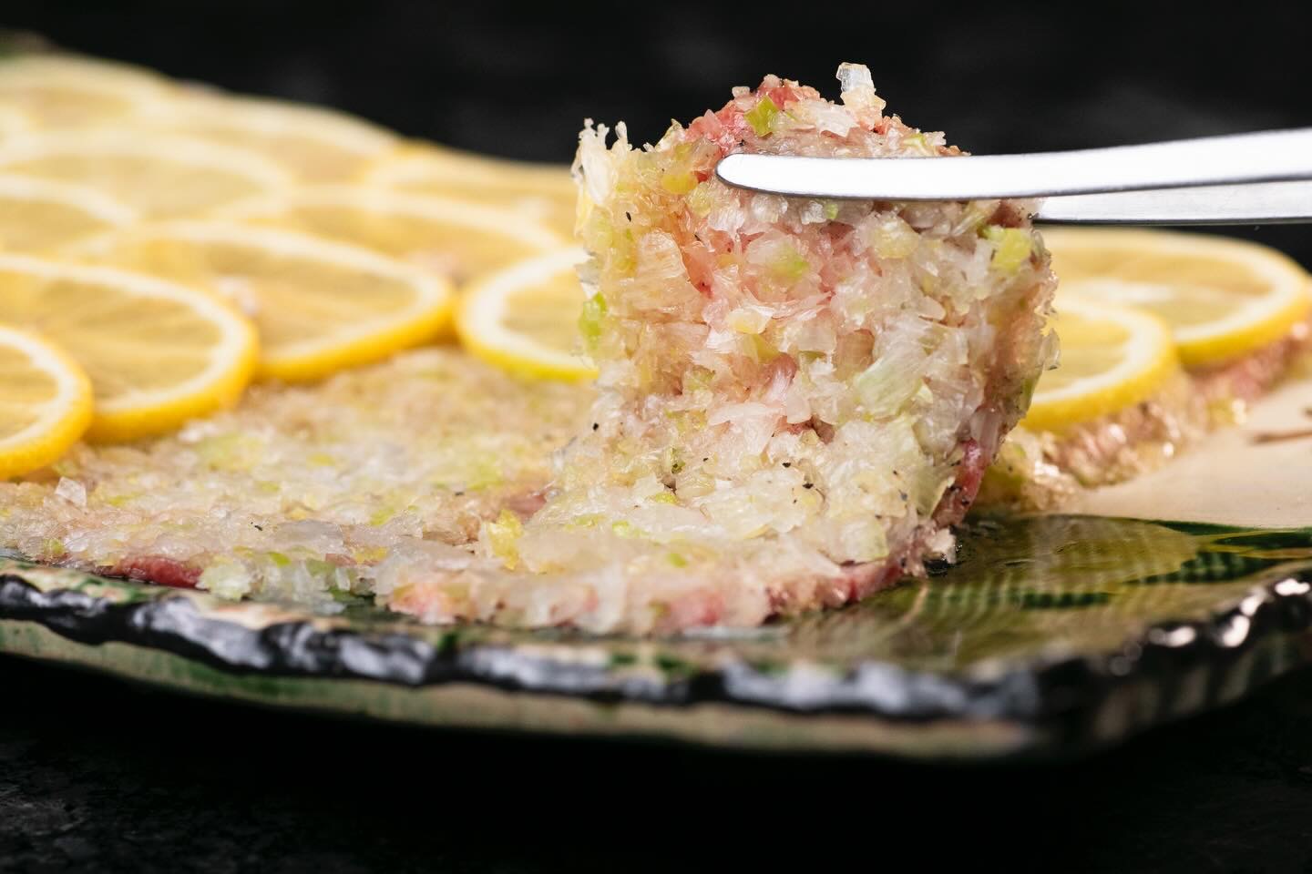 レモンの輪切りを並べた皿から、刻みねぎと黒こしょうをのせた薄い肉をトングで持ち上げたところ。